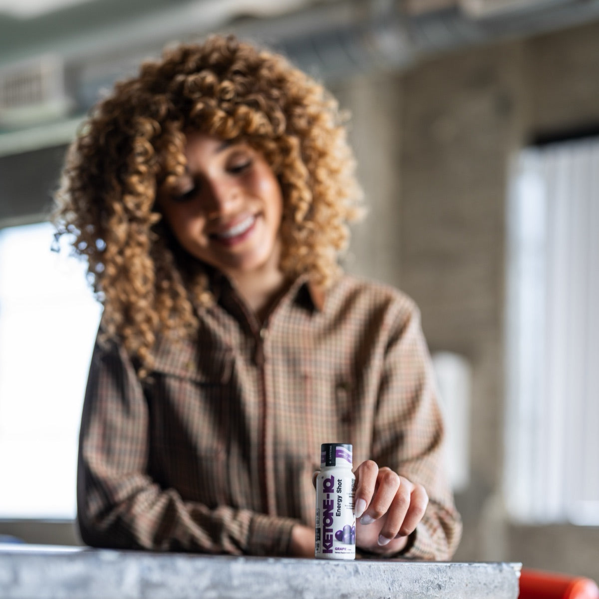 Smiling woman with curly hair resting a Ketone-IQ +Caffeine Grape shot on a counter
